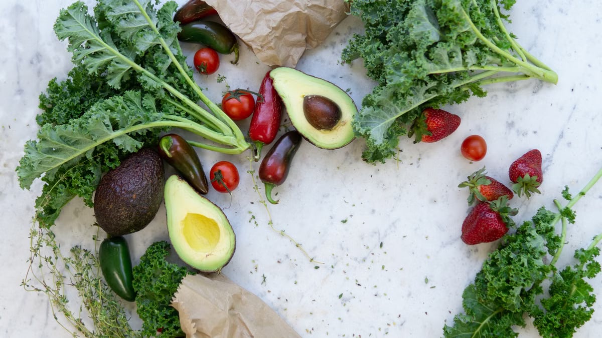 Top view of fresh avocados, kale, tomatoes, and peppers arranged on a marble surface.